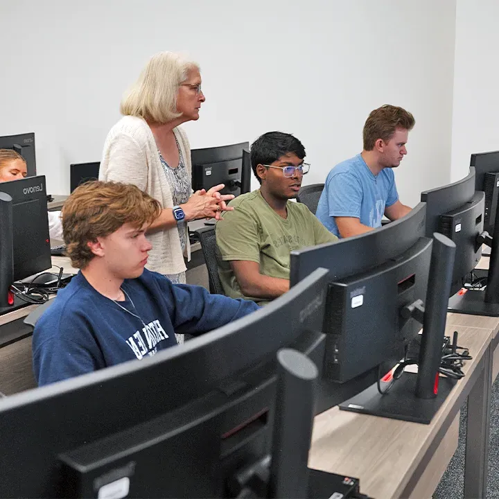 Three students working on desktop computers while professor looks on.